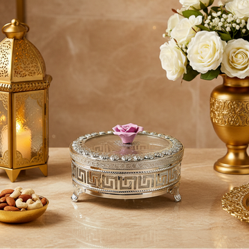 An ornate oval silver-toned jewelry box with a clear glass lid, decorated with a Greek key pattern and small feet, topped with a pink rose on a black background.